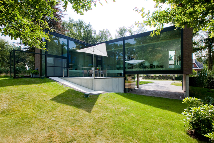 le côté d'une maison est fait de verre et d'aluminium. Il y a de l'herbe à l'avant de l'image et des arbres de chaque côté. C'est une journée ensoleillée.