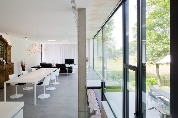 an internal shot of a dining room. There is a glass and aluminium wall to one side which is letting in plenty of natural light. A white dining table with some chairs is on the other side of the room.